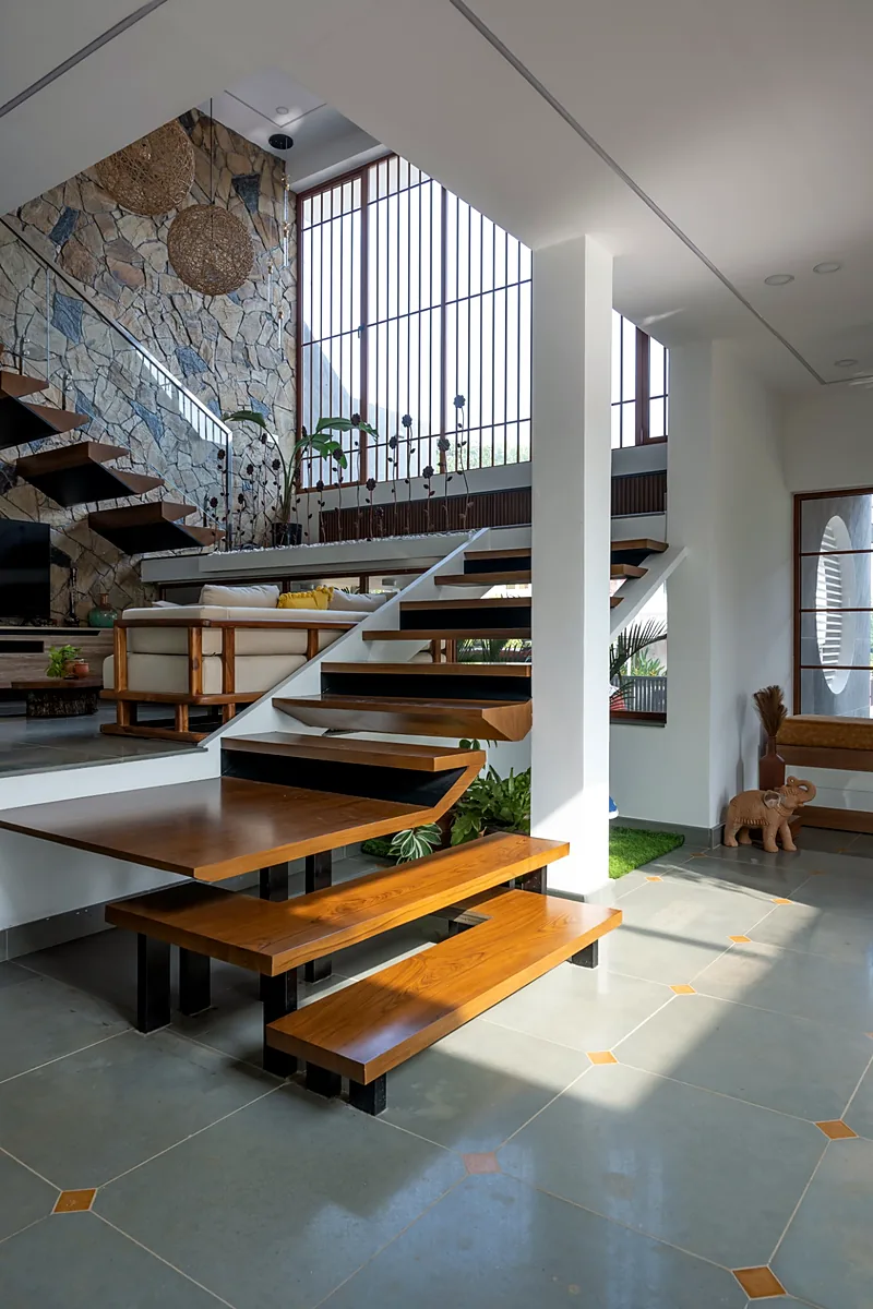 Double-height living room with a floating wooden staircase, stone wall and tall louvred window in a modern tropical house in Kerala